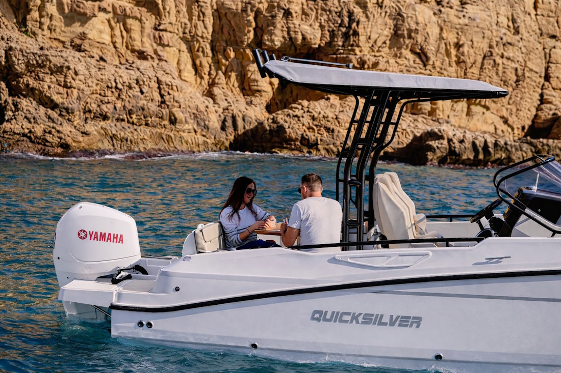 Couple having drinks on the stern deck of a Quicksilver boat anchored next to golden rocky cliffs